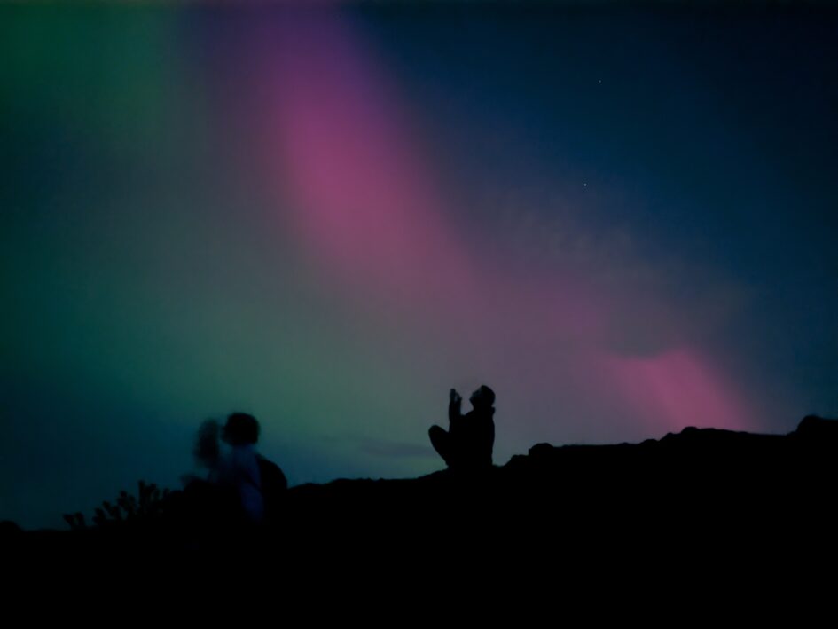 Image shows a woman sitting looking up at the sky and taking photos with her phone. She is silhouetted against the sky which is green and purple